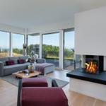 Ein Wohnzimmer mit Kamin, grauem Sofa und roten Sitzkissen. Große Fenster bieten Blick auf die Landschaft.