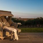 Rattan-Sonnenschutz mit Tisch und Getränken auf Holzterrasse. Blick auf Grünfläche und Horizont.