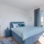Schlafzimmer mit blauer Bettwäsche und großem Fenster zum Balkon.