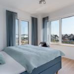Schlafzimmer mit Bett, Fenstern und Blick auf Wohnhäuser.