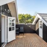 Deck with wooden floor, chair, and table. Black wood walls and white doors. View of trees.