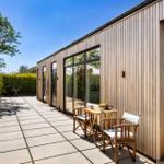 Terrace with wooden table and chairs beside modern wooden house with large windows.