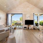 Living area with sofa, armchairs, TV, and dining area. Wooden walls and large windows.