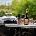 Closed grill with beer, glasses, and spice on table outdoors.