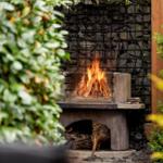 Outdoor area with burning wood stove, surrounded by plants and stone wall.