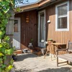 House with wooden siding, patio, table, chair, and plants in the front yard.