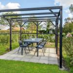 Black pergola with table and chairs on stone patio in garden with grass and shrubs.