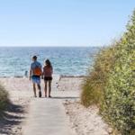 Two people walk along a path leading to the beach. Bushes grow on both sides.