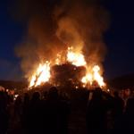 Großer Scheiterhaufen brennt bei Nacht. Menschen beobachten das Feuer.