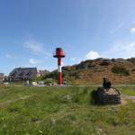 Rotes Leuchtturm mit weißer Markierung, Grasland und Häuser im Hintergrund.