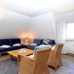 Wohnzimmer mit blauem Sofa, Rattanstühlen und Holztisch. Fenster mit weißen Vorhängen.