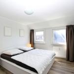 Schlafzimmer mit Doppelbett, zwei Fenstern und Nachttisch mit Lampe.