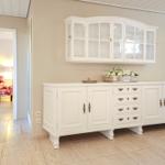 Weißer Sideboard mit Schubladen und darüber hängender Schrank. Im Hintergrund ist ein roter Sessel sichtbar.