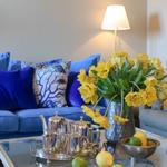 Blauer Sofa mit Kissen, Glas-Tisch mit Teeservice und gelben Tulpen.