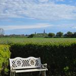 Eine weiße Bank steht im Garten vor einem Heckenzaun. Im Hintergrund sind Felder und ein Dorf mit Kirchturm zu sehen.