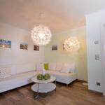 Wohnzimmer mit weißem Sofa, grünen Kissen und runden Tisch. Zwei Doldenlampen hängen von der Decke.