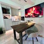 Essbereich mit Holztisch, zwei Stühlen und Küchenbereich. Wandfernseher zeigt Flammen.