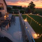 Terrasse mit Tisch, Stühlen und Lichterkette bei Sonnenuntergang. Grünfläche und Garten im Hintergrund.