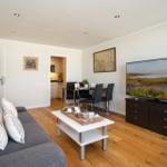 Wohnzimmer mit Sofa, Esstisch und Fernseher. Holzdielenboden und offene Küche im Hintergrund.