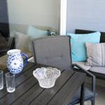 Terrasse mit Tisch, Stühlen und Kissen. Auf dem Tisch steht eine Karaffe und Gläser.