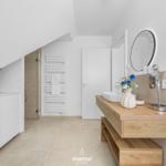 Bathroom with wooden vanity, round mirror, and shower under sloped ceiling.
