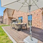 Deck with table, chairs, and umbrella next to a brick house with thatched roof.