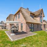 House with thatched roof and wooden terrace with table and umbrella.