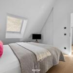 Schlafzimmer mit Dachschräge, Bett, Fenster und TV. Durchgang zu Wohnbereich mit Stuhl.