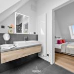 Badezimmer mit Waschtisch und Spiegel. Im Hintergrund ist ein Schlafzimmer mit Bett und Dachfenster.