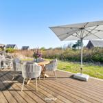 Terrasse mit Tisch, Stühlen und Sonnenschirm. Hintergrund: Gras, Sträucher und Häuser.