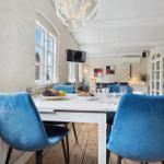 Dining area with blue chairs and white table. Wooden floor and windows visible.