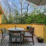 Terrace with table, chairs, and grill. Yellow stucco and wooden decking. View into green trees.