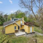Yellow house with terrace and garden. Surrounded by trees under blue sky.