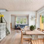Dining area with wooden table and chairs, view into living room with sofa and windows.