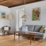 Living room with gray sofa, wooden floor, and ceiling beams. Two paintings hang on the wall.