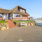 House with thatched roof, stone wall, and terrace with white bench.