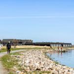Wohnhäuser am Wasser mit Windrad im Hintergrund. Menschen spazieren entlang des Ufers.