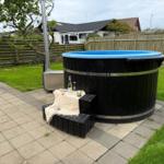 Schwarzer Holzhot tub mit blauem Rand auf gepflastertem Hof, umgeben von Gras und Holzzaun.