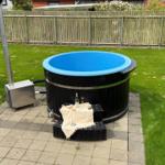 Ein schwarzer Whirlpool mit blauem Innenbereich steht auf einer Terrasse mit Grünfläche.