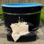 Ein schwarzer Holzwhirlpool mit blauem Innenrand. Davor steht eine Bank mit Tischdecke, Wein, Glas und Kerze.