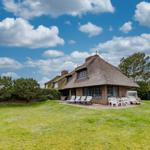 Haus mit Strohdach, Terrasse mit Liegen und Tisch, umgeben von Gras und Bäumen unter blauem Himmel.