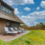Haus mit Strohdach, Terrasse mit Liegestühlen und grünem Rasen unter blauem Himmel.