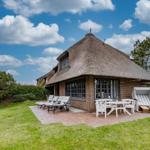 Dorfhaus mit Strohdach, Terrasse mit Tisch, Stühlen und Liegestühlen auf grünem Rasen.
