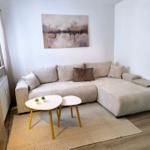 Wohnzimmer mit Ecksofa, zwei Tischen und Wandbild.