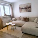 Wohnzimmer mit Ecksofa, zwei Tischen und Wandbild. Fenster mit Jalousien und Vorhängen.