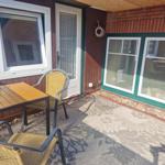 Terrasse mit Tisch, zwei Stühlen und Teppich. Hintergrund: Hauswand mit Fenstern und Tür.