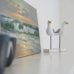 Zwei hölzerne Möwenfiguren stehen auf einem weißen Tisch neben einem Strandgemälde.