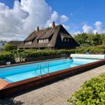 Pool mit Holzrand und Schwimmsteg vor einem Dachhaus mit Strohdach.
