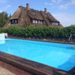 Ein Schwimmbad mit Holzrand und Steinmauer. Im Hintergrund ein Dachstuhlhaus mit Strohdach.