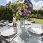 Garten-Tisch mit Blumen, Gläsern und Platzsetzung. Hintergrund: Grünfläche und Haus.